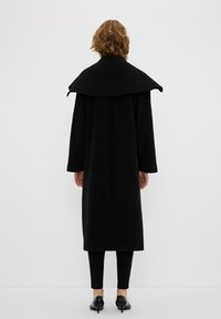 Cappotto di lana nero con un colletto oversize a ribalta, maniche lunghe e silhouette dritta; indossato sopra leggings neri e scarpe con tacco.