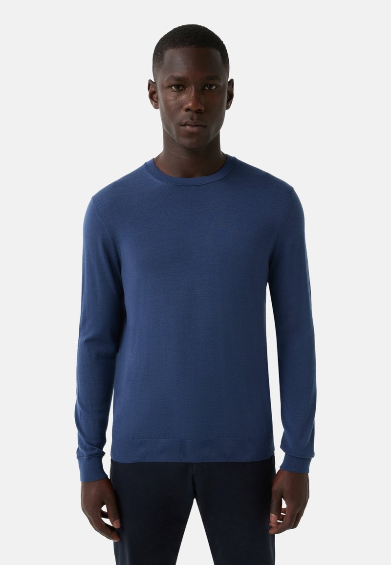 Mężczyzna ubrany w prostą, niebieską bluzę z długim rękawem i okrągłym dekoltem, stojący na białym tle, zwrócony w przód z neutralnym wyrazem twarzy.