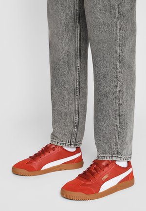 Osoba ubrana w szare dżinsy i czerwone buty sportowe Puma z białym paskiem i gumową podeszwą, stojąca na jednolitym jasnym tle.