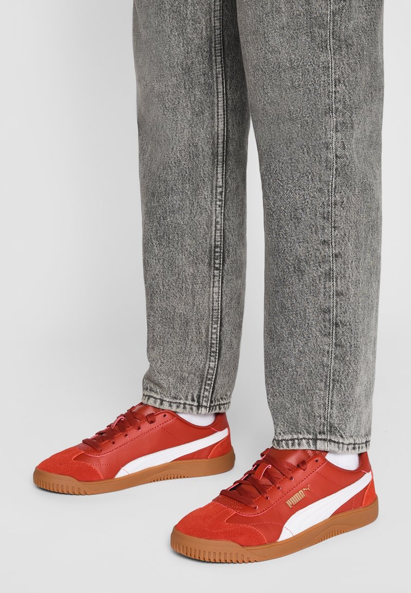 Personne portant un jean gris et des baskets rouges Puma avec une bande blanche et une semelle en gomme, debout sur un fond clair uni.