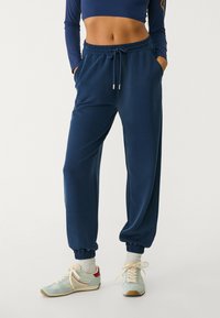 Dunkelblaue Sweatpants mit elastischem Bund, umschlagenen Knöcheln, Seitentaschen und einer glatten Textur, kombiniert mit hellblauen Sneakern.