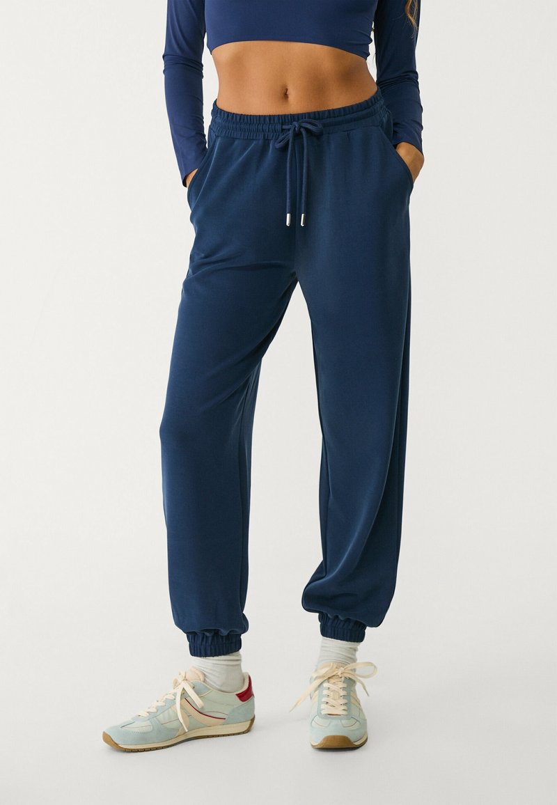 Dunkelblaue Sweatpants mit elastischem Bund, umschlagenen Knöcheln, Seitentaschen und einer glatten Textur, kombiniert mit hellblauen Sneakern.
