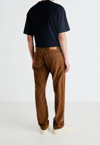 Hnědé kordové kalhoty s zadními kapsami a koženou nášivkou na pase, spárované s černým tričkem s krátkým rukávem a bílými teniskami.