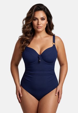 Kobieta z długimi, falującymi włosami, ubrana w granatowy jednoczęściowy kostium kąpielowy z detalami w postaci złotych pierścieni na ramiączkach, stojąca na tle jednolitego białego tła.