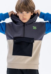 Sweatshirt à capuche composé d'un mélange de matériaux gris, bleu et beige. L'avant comporte une grande poche noire avec des boutons-pression. Texture douce.