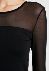 Černý top s průhlednou síťovanou horní částí a dlouhými rukávy, doplněný hladkou, neprůhlednou dolní částí. Jednoduchý, moderní design.