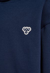 Mörkblå tröja med ribbad textur; har en kontrasterande broderad logotyp i vitt. Mjuk, elastisk tyg; minimalistisk design.