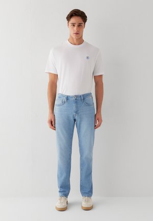 Junger Mann steht frontal, trägt ein einfaches weißes T-Shirt, hellblaue Jeans und beige Sneakers vor einer weißen Wand.
