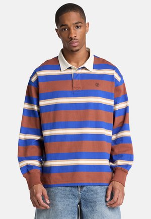 Giovane uomo che indossa una polo a maniche lunghe con ampie strisce orizzontali blu, marrone e crema, abbinata a jeans azzurri.