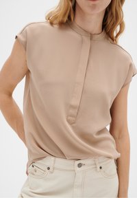 Beige blus med kort ärm, rund halsringning och nyckelhållsknappar. Slät textur, avslappnad passform, kombinerad med ljusa jeans.