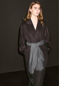 Long black coat with a wide collar, oversized fit, and a gray knitted belt. Smooth texture and minimalist design enhance modern appeal.