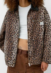 Kurtka w nadruk w panterę z czarnym skórzanym kołnierzem, zamek błyskawiczny, kieszenie z przodu i mała metka z logo, połączona z białą bluzką i brązowymi spodniami.