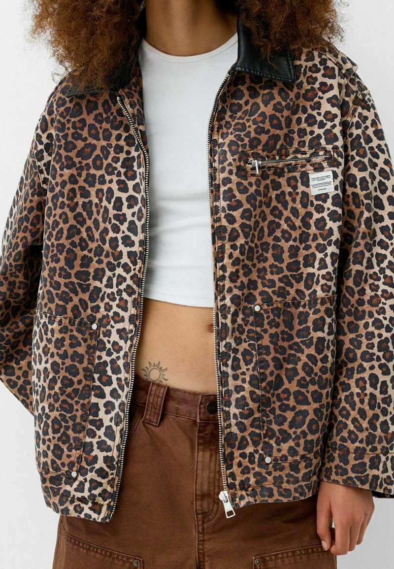 Kurtka w nadruk w panterę z czarnym skórzanym kołnierzem, zamek błyskawiczny, kieszenie z przodu i mała metka z logo, połączona z białą bluzką i brązowymi spodniami.