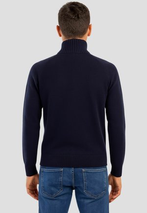 Maglione in maglia blu navy con collo alto a coste, design aderente e polsini e orlo a coste. Indossato con jeans blu.