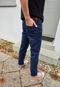 Osoba v tmavě modrých džínách a béžových teniskách stojí venku na dlážděné ploše poblíž bílých schodů a kovových roletových dveří.