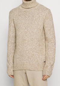 Pull col roulé beige en tissu tricoté texturé, avec un col et des poignets côtelés, une coupe décontractée et une petite étiquette sur le côté.