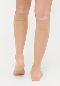 Beige kniekousen met een textuurband aan de bovenkant, doorschijnend materiaal en een gladde afwerking, gezien van achteren.