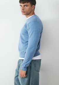 Jasnoniebieski sweter z okrągłym dekoltem, z długimi rękawami, z ribbingu przy mankietach, o miękkiej fakturze; noszony na białej koszuli i w zestawie z dżinsami.