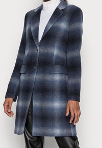 Vrouw in een blauw-grijs geblokt jasje met een gradient, over een witte coltrui en zwarte broek, staande tegen een eenvoudige witte achtergrond.