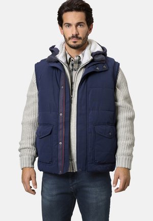 Marineblaue Steppweste mit Kapuze, ausgestattet mit einem fleecegefütterten Kragen, vorderen Taschen und kontrastierenden Nähten. Getragen über einem grauen strukturierten Pullover.