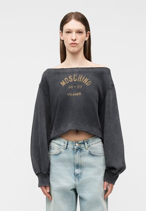 Žena v čiernej vyblednutej mikine s odhalenými ramenami s textom "Moschino Jeans 86 23" a svetlomodrých džínsach s vysokým pásom, stojaca pred jednoduchým pozadím.