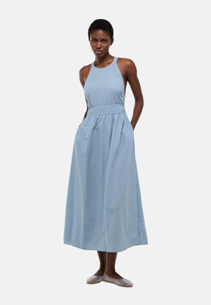 Femme portant une robe sans manches bleu clair, longue jusqu'aux chevilles, avec un col haut et des poches, associée à des chaussures plates transparentes à enfiler.