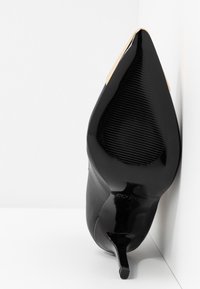 Czarny szpilka z ostrym noskiem, z teksturowaną podeszwą i złotym metalowym akcentem na końcu, ustawiona w pionie przy białym kącie.