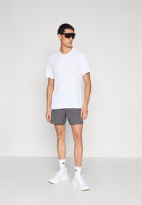 Camiseta deportiva blanca, pantalones cortos grises, zapatillas blancas y gafas de sol oscuras; diseño minimalista con un pequeño logo en el pecho izquierdo de la camiseta.