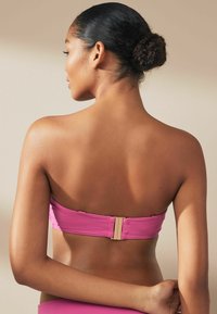 Top bikini rosa senza spalline con chiusura dorata sul retro, realizzato in un tessuto morbido e con un design aderente che definisce le spalle.