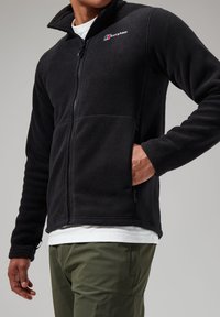Black fleece jacket with a high collar and zip closure, featuring a logo on the left chest and side pockets. Worn over a white shirt.