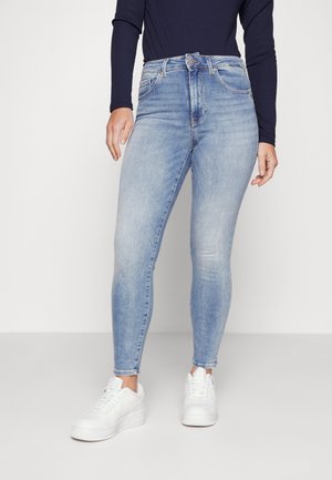 Jean skinny bleu clair en denim, avec une taille mi-haute, un design classique à cinq poches et une texture légèrement délavée. Associé à des baskets blanches.
