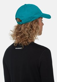 Teal katoenen pet met een gebogen bril en een verstelbare strap aan de achterkant, gedragen met een zwart shirt met lange mouwen met een wit logo.