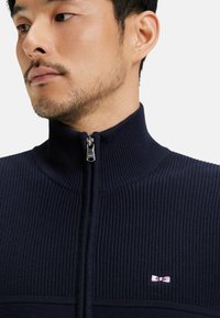 Pull-over zippé bleu marine avec une texture côtelée, un col montant et un petit logo rose sur la poitrine. Équipé d'une fermeture éclair en métal argenté.