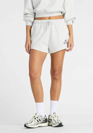 Person trägt hellgraue New Balance Shorts, passenden Pullover, weiße Sportsocken und silberne Laufschuhe und steht vor einem einfarbigen Hintergrund.