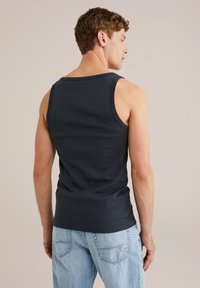 Hombre con una camiseta sin mangas negra y jeans azul claro, de pie con la espalda hacia la cámara contra un fondo neutro.