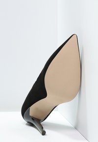 Svart spetsig högklackad sko visad från botten, med beige sula och smal klack, placerad upprätt mot vita hörnväggar.