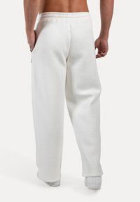 Homme portant un pantalon blanc ample et des baskets blanches, se tenant avec les mains détendues sur les côtés, vu de dos sur un fond blanc.