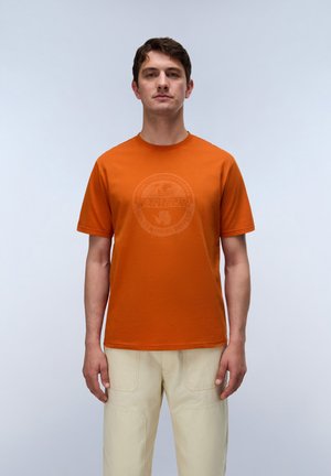 Mand står iført en orange kortærmet t-shirt med svagt logo og cremefarvede bukser mod en lys ensfarvet baggrund.