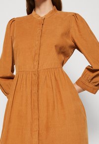 Vestito in velluto a coste di un caldo arancione, caratterizzato da un colletto alto, chiusura con bottoni, maniche a sbuffo e una gonna plissettata per texture e forma.