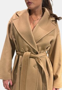 Cappotto doppiopetto beige realizzato in tessuto strutturato, con ampi revers, cintura e grandi bottoni. Finitura liscia con tasche nascoste.