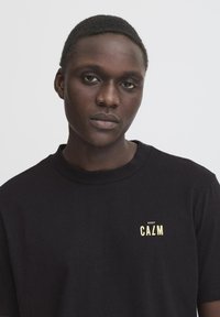 Svart bomull t-skjorte med rund hals og korte ermer, med et lite "KEEP CALM"-grafikk i gult på brystet.