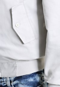 Zbliżenie na kieszeń białej kurtki z detalem guzika nad pasem wyblakłych niebieskich dżinsów.