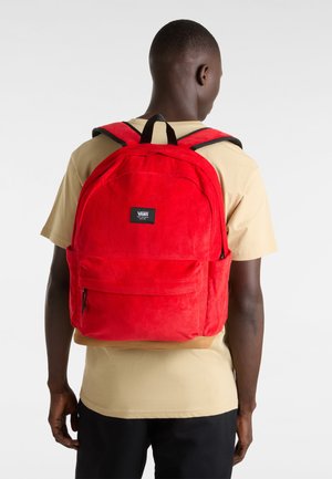 Mochila de pana roja con dos bolsillos delanteros. Cuenta con tirantes y detalles negros, forma rectangular y un parche con el logotipo en la parte frontal.