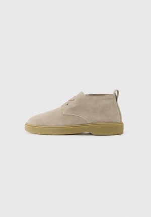 Zapato de ante beige de altura hasta el tobillo, con cordones y suela de goma marrón claro, con tira en la parte trasera del talón, logo de Calvin Klein cerca del cuello.