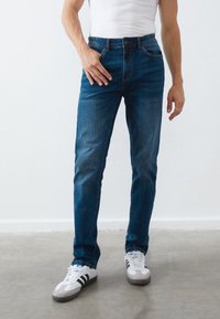 Jeans in denim blu scuro, dal taglio slim, con cinque tasche, chiusura a bottoni e zip, e un leggero effetto sbiadito. Indossati con scarpe da ginnastica bianche.