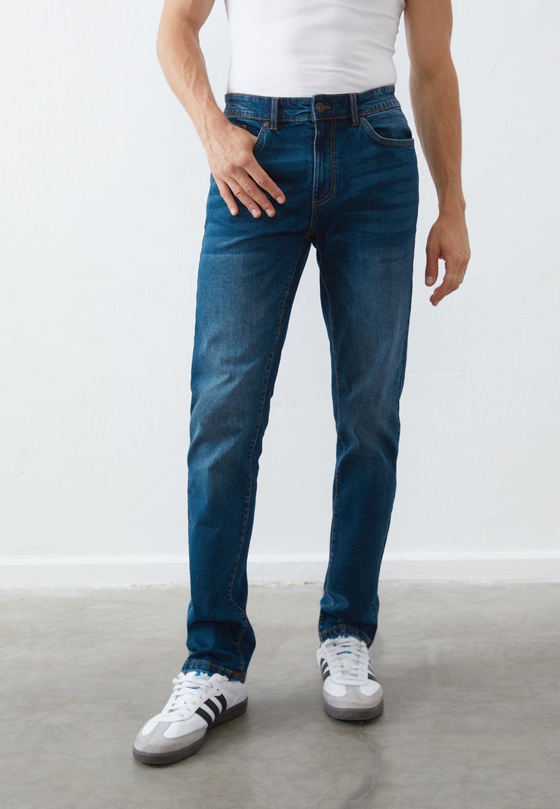 Dark blue denim jeans with a slim fit, featuring five pockets, a button and zipper closure, and subtle fading. Worn with white sneakers.