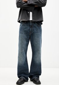 Helle blaue Jeans mit lockerer Passform, die ein Fünf-Taschen-Design, dezente Ausbleichungen und gestickte Akzente aufweisen. Kombiniert mit schwarzen Schuhen.