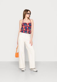 Top in tessuto floreale rosso con foglie blu, abbinato a pantaloni bianchi a vita alta e a gamba larga. Accessoriato con una borsa arancione e occhiali da sole.