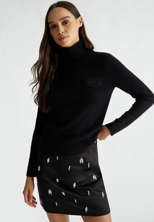 Maglione nero con colletto alto e polsini a coste, dotato di una tasca patch testurizzata. Indossato con una gonna nera lucida e decorata.