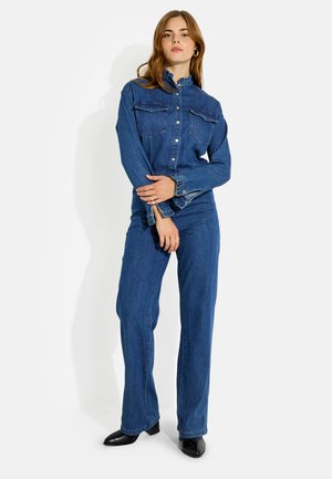 Mujer con cabello largo, vestida con una camisa vaquera azul de botones y vaqueros de pierna ancha a juego, de pie con los brazos cruzados a la altura de la cintura.